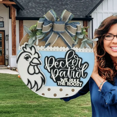 A smiling woman with glasses holds a decorative round sign featuring a chicken illustration and the words Pecker Patrol, We Rule The Roost, standing in front of a modern house with a wooden front door.