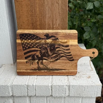 A wooden cutting board with an engraved image of a cowboy riding a bucking horse in front of a waving American flag, resting on a white brick surface with greenery in the background.