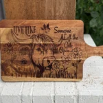 A wooden cutting board engraved with a cow, flowers, a bird, and the phrase “Live like someone left the gate open,” resting on a white brick ledge with greenery in the background.