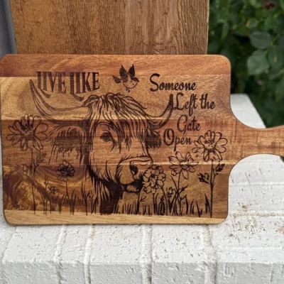 A wooden cutting board engraved with a cow, flowers, a bird, and the phrase “Live like someone left the gate open,” resting on a white brick ledge with greenery in the background.