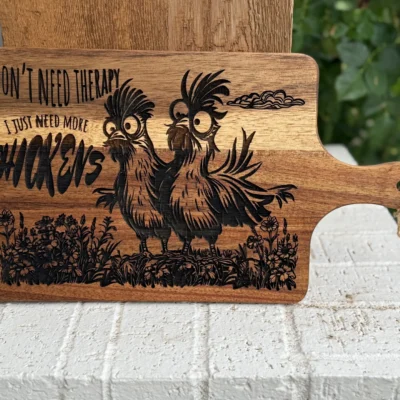 A wooden cutting board features a drawing of two quirky chickens and the text, I dont need therapy, I just need more chickens, engraved above and next to the birds. The board is outdoors, propped on a white brick surface.
