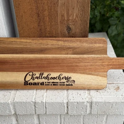 A wooden cutting board with the words Chattahoochee Board: a lot about livin and a little bout lunch engraved on it, rests on a white brick ledge with greenery in the background.