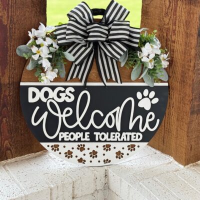 A round sign with a black and white striped bow, flowers, and greenery reads, Dogs welcome, people tolerated, with paw prints, hanging on a wooden wall.
