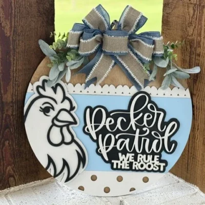 A round wooden sign with a drawing of a chicken, a large blue and burlap bow, greenery, and the text “Pecker Patrol We Rule The Roost” in white and black lettering.